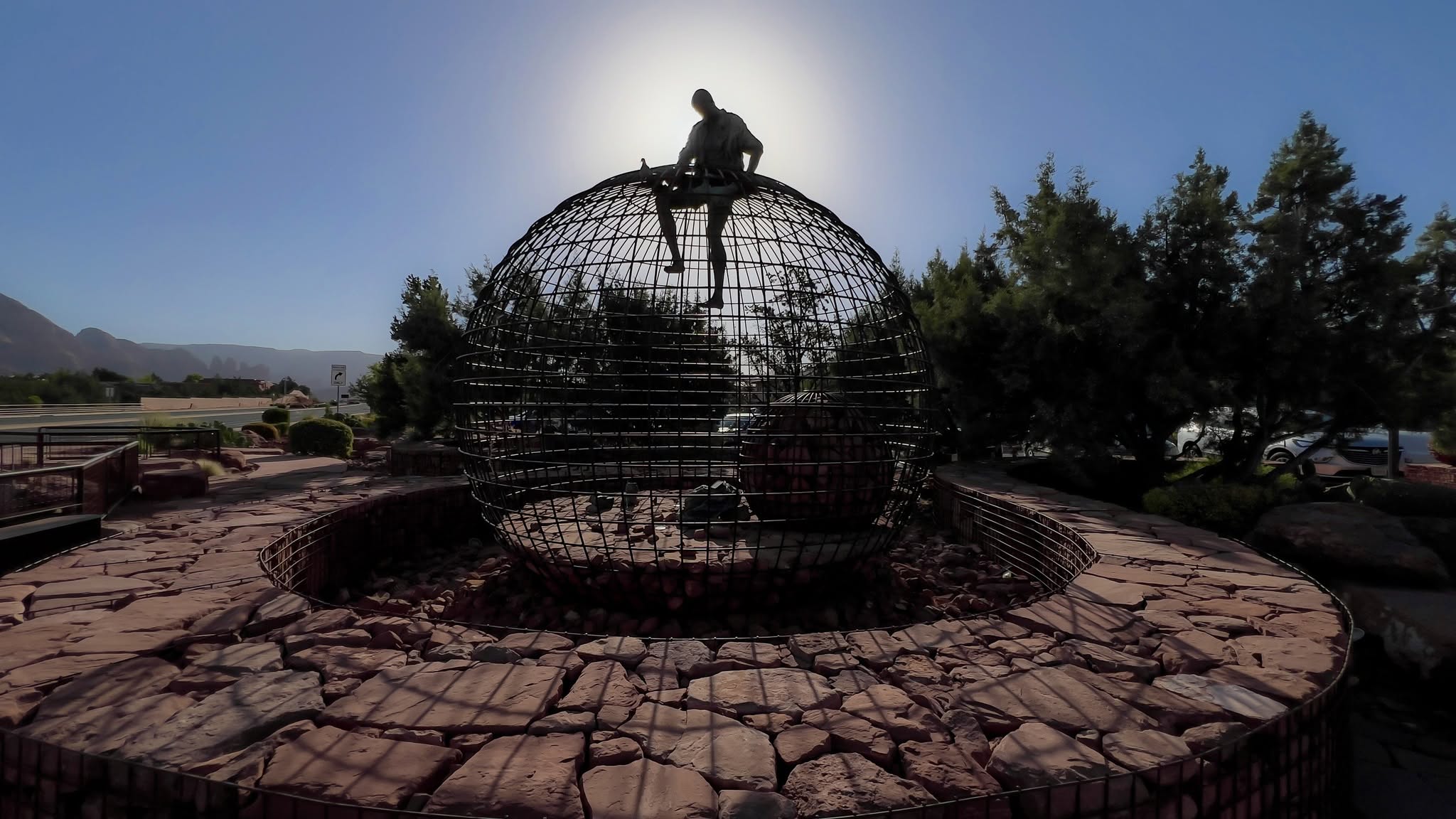 Silhouette of a figure climbing out of a steel globe sculpture against the sun
